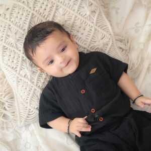 Charming baby dressed in black, smiling on a textured cushion indoors.