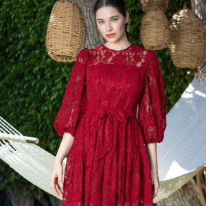 Stylish woman in red lace dress standing outdoors with wicker baskets and hammock.