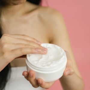 Close-up of a woman applying white cream from a jar against a pink background.